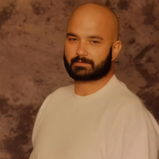 Portrait of Kris, a bald man with a dark beard, wearing a white shirt against a textured brown background.