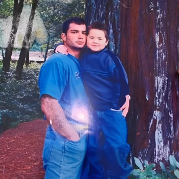 A man and a young boy stand together in a forest, leaning against a large tree trunk.