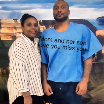 A man and a woman standing together in front of a canyon backdrop. The man wears a blue t-shirt with the text 'Mom and her son love you miss you'.