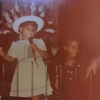Victor3, a young boy in a white hat and dress, sings into a microphone on a stage.