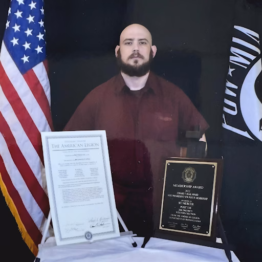 A portrait of Travis, 39, with an American Legion certificate and a membership award plaque displayed in front of him.