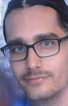 Daniel Close-up portrait of Daniel, a man with dark hair, wearing black-framed glasses and a light beard.
