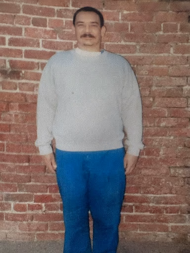A man with a mustache wearing a light grey sweater and blue pants stands against a red brick wall.