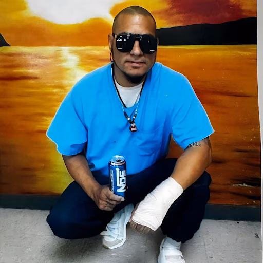 Giovanny crouching in front of a sunset mural, wearing a blue shirt, sunglasses, and a bandaged hand, holding a beer can.
