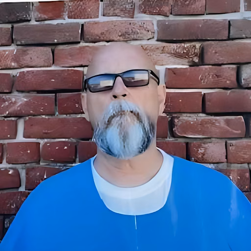 James, a bald man with a white goatee and glasses, wearing a blue shirt, stands in front of a red brick wall.