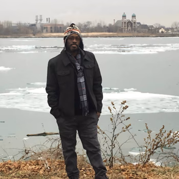 Marcus stands on a frozen shoreline in winter, wearing a beanie and scarf, with a church and industrial buildings in the background.