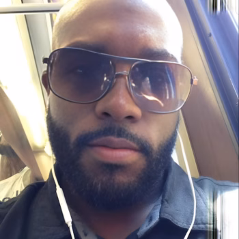 Marcus, a bald man with a beard and sunglasses, wearing white earphones, looks directly at the camera.