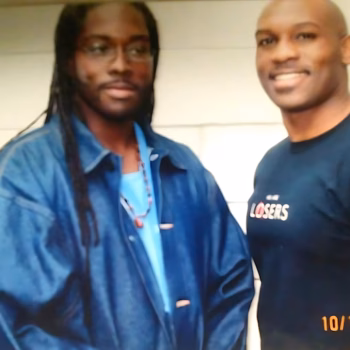 Marcus and Ja'far, two men standing together. Marcus wears a blue jacket over a light blue shirt and glasses, while Ja'far wears a dark blue t-shirt with the word 'LOSERS' printed on it.