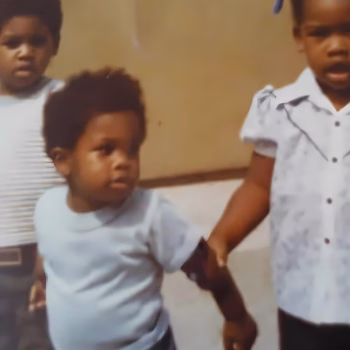 Three young children, Marcus, Ja'far, and another, are standing together indoors.
