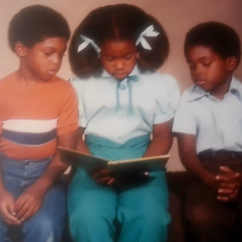 Three young Black children in school uniforms sit together, looking down at an open book.