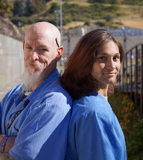 A bald man with a white beard and a woman with brown hair, both wearing blue scrubs, stand back-to-back outdoors.