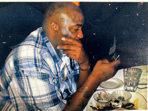 A man in a plaid shirt sits at a table, looking down at a phone in his hand.