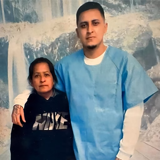 A man in a blue surgical gown stands with his arm around an older woman in front of a waterfall backdrop.