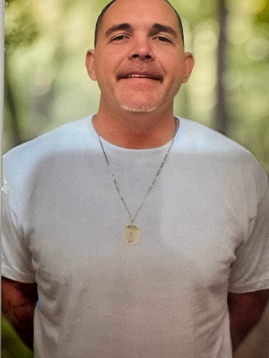 Bradley, 47, a man with a mustache and goatee, wearing a white t-shirt and a necklace, smiles at the camera against a blurred green background.