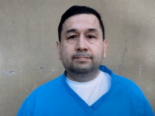 Edgar, a man with dark hair and a light beard, wearing a blue V-neck shirt over a white undershirt, against a plain beige wall.