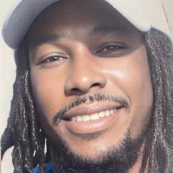 Close-up portrait of Jasper, a man with dreadlocks, a beard, and a cap, smiling at the camera.