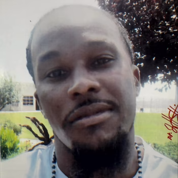 A close-up portrait of Jasper, a man with a beard and a beaded necklace, outdoors with a building and trees in the background.