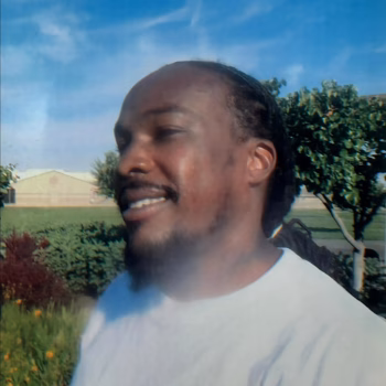 A man with braided hair and a beard, wearing a white t-shirt, smiles outdoors under a blue sky.
