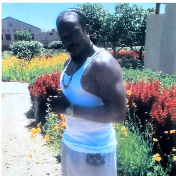 Jasper, 43, stands in a sunlit garden filled with red and yellow flowers, wearing a white tank top and a decorative belt.
