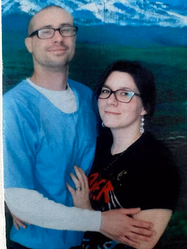 A man and a woman wearing glasses stand together in front of a mountain landscape backdrop.