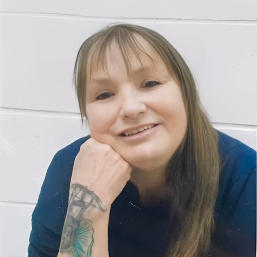 Portrait of Melonie, a woman with long brown hair and a tattoo on her wrist, resting her chin on her hand against a white wall.