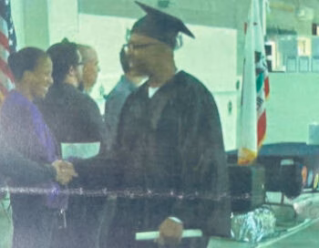 Norman4, a man in a graduation cap and gown, shakes hands with a woman in a purple suit at an indoor event.