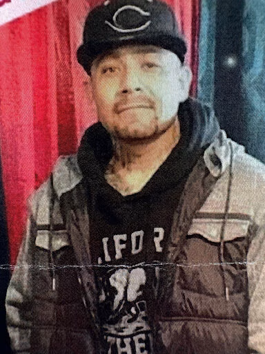A man identified as Amado, 47, wearing a Chicago Cubs cap, a black hoodie, and a puffer vest, standing in front of a red curtain.