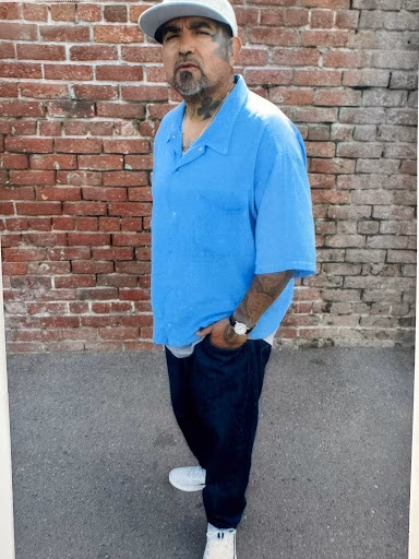 Tony4, a man with a goatee and tattoos, stands in front of a red brick wall wearing a blue shirt, dark pants, and a white cap.