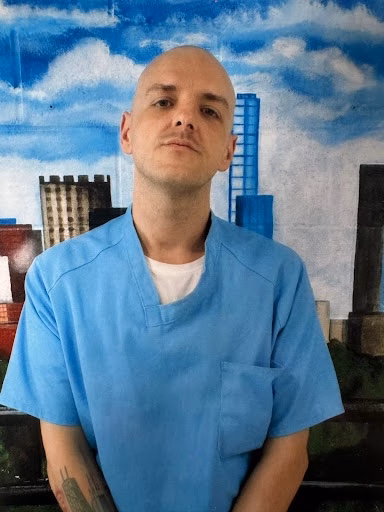 A bald man in blue scrubs stands before a painted cityscape mural.