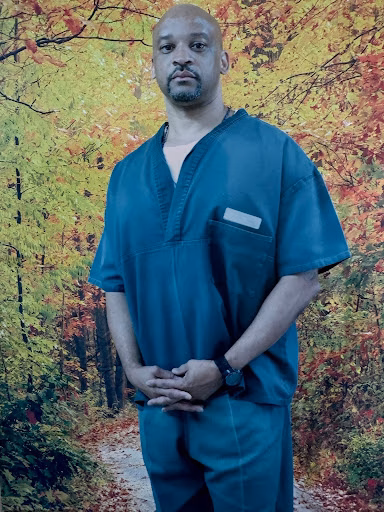 A man in teal scrubs stands against a backdrop of a vibrant autumn forest.