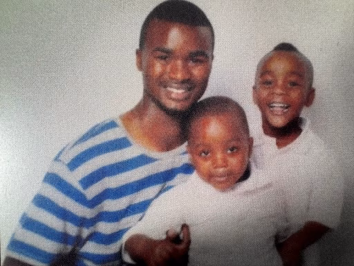 A smiling man in a blue and white striped shirt holding two young children against a plain background.
