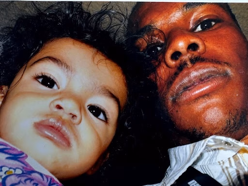 A close-up photograph of a man and a young child lying together, looking directly at the camera.