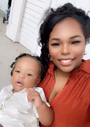 A woman with dark curly hair in a red shirt holds a young child with braided hair in a white shirt, both looking at the camera.