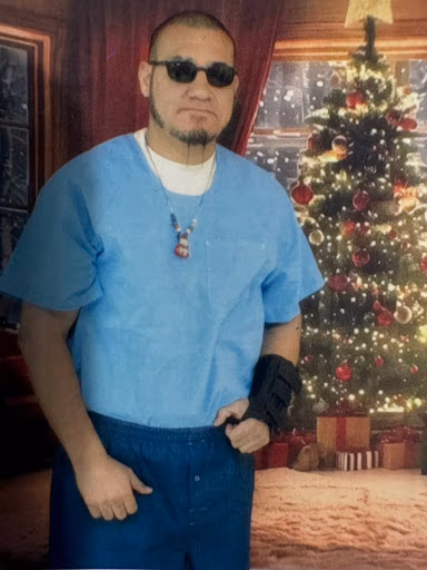 Giovanny in blue scrubs and sunglasses stands before a decorated Christmas tree.