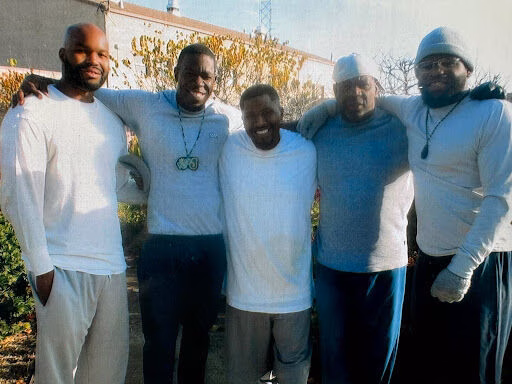 Five men stand together outdoors, smiling and with arms around each other's shoulders.
