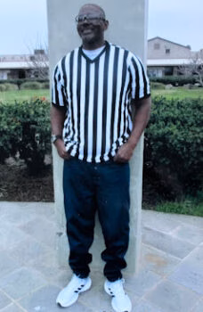 Jonathan, a man in a black and white striped referee shirt and dark pants, stands smiling with hands in pockets.