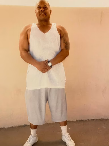 Keith, a bald man in his 50s, stands against a plain wall wearing a white tank top, grey shorts, and white sneakers.