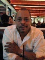 Keith, 57, a man with a shaved head and a white shirt, sits with his arms crossed in a restaurant.