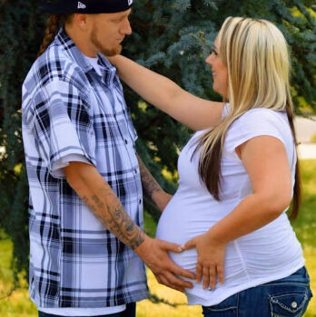 A pregnant woman in a white shirt and jeans stands next to a man in a plaid shirt, both looking at her baby bump.