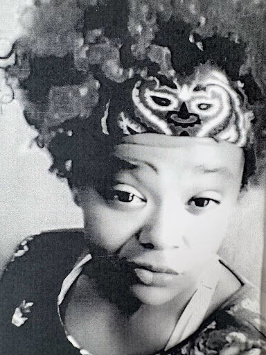 Black and white portrait of Zoe, a woman with a large afro hairstyle and a decorative mask headband, looking directly at the camera.