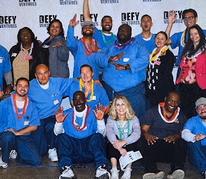 A diverse group of people in blue scrubs and lanyards posing for a group photo in front of a Defy Ventures backdrop.