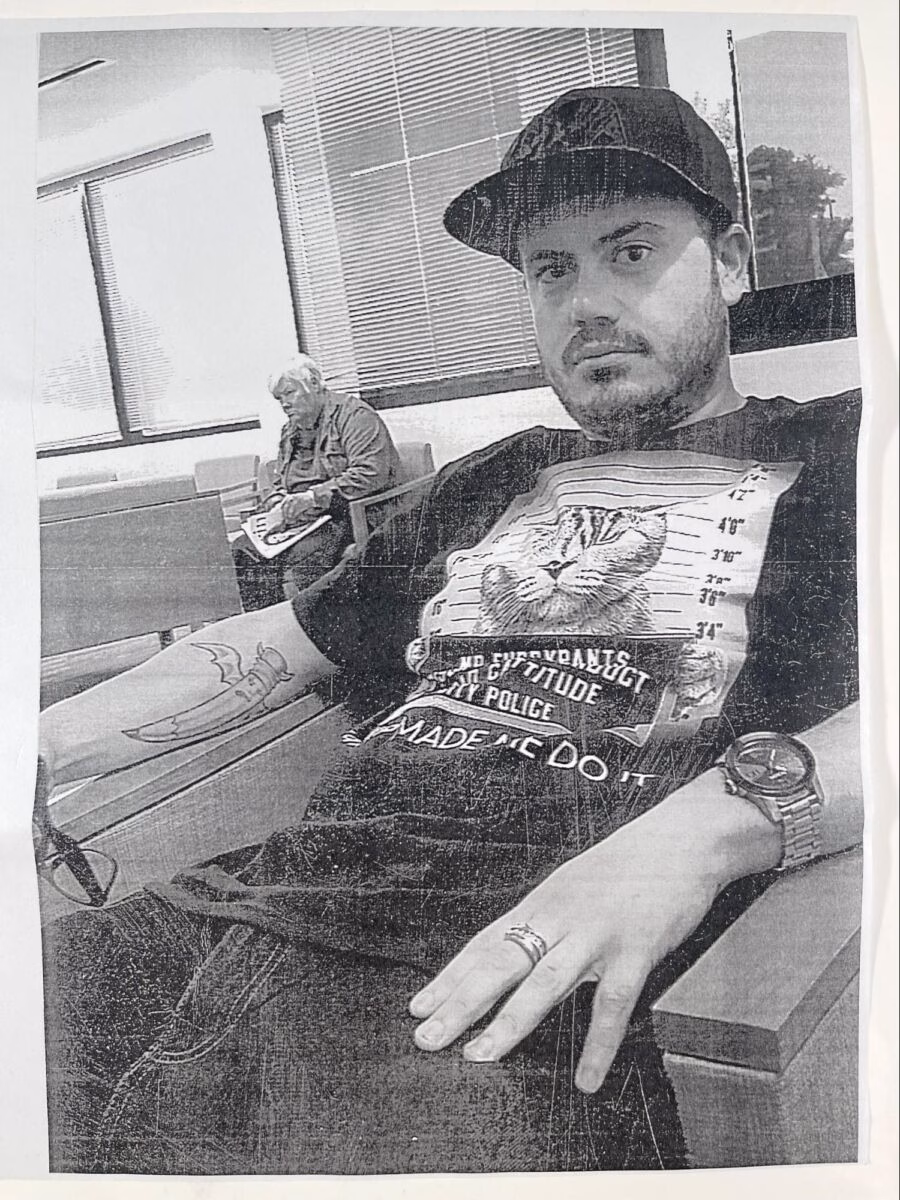 A man wearing a baseball cap and a graphic t-shirt sits in a waiting area, with another person visible in the background.