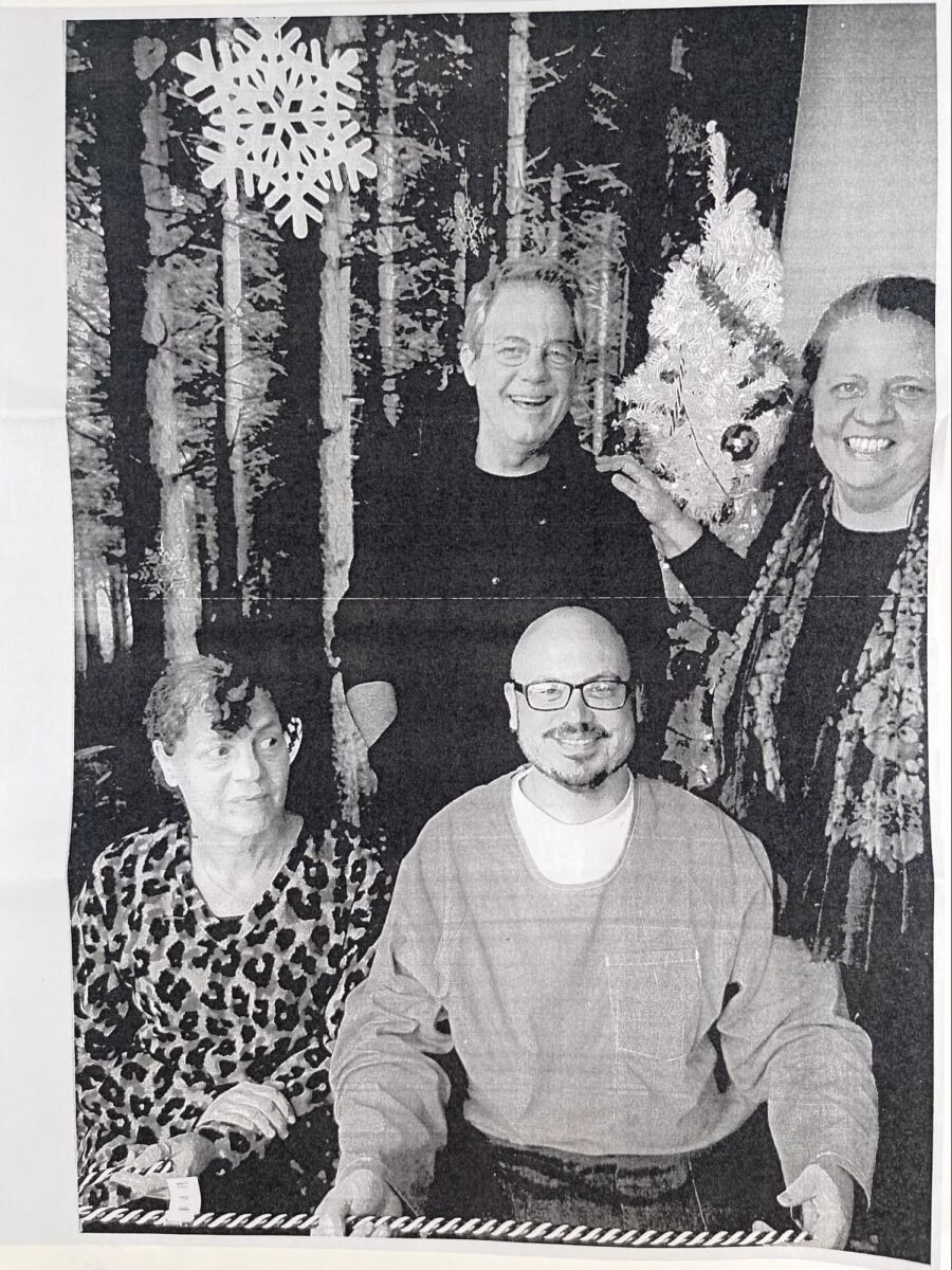 Four people posing for a photo in front of a forest backdrop with a large snowflake and a small Christmas tree.