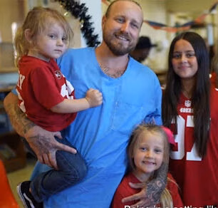 A man in a blue shirt holds a young child in a red shirt, with two other children in red jerseys standing nearby.