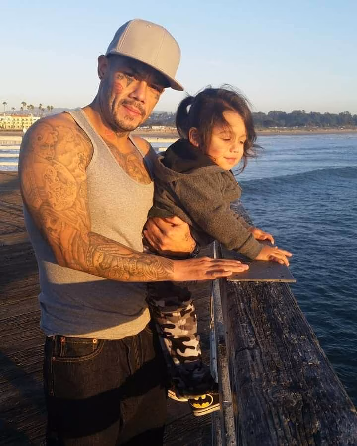 A man with tattoos and a cap holds a young child on a wooden pier overlooking the ocean.