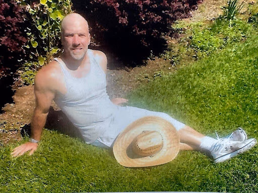 A bald man in a white tank top and shorts sits on a green lawn with a straw hat beside him.