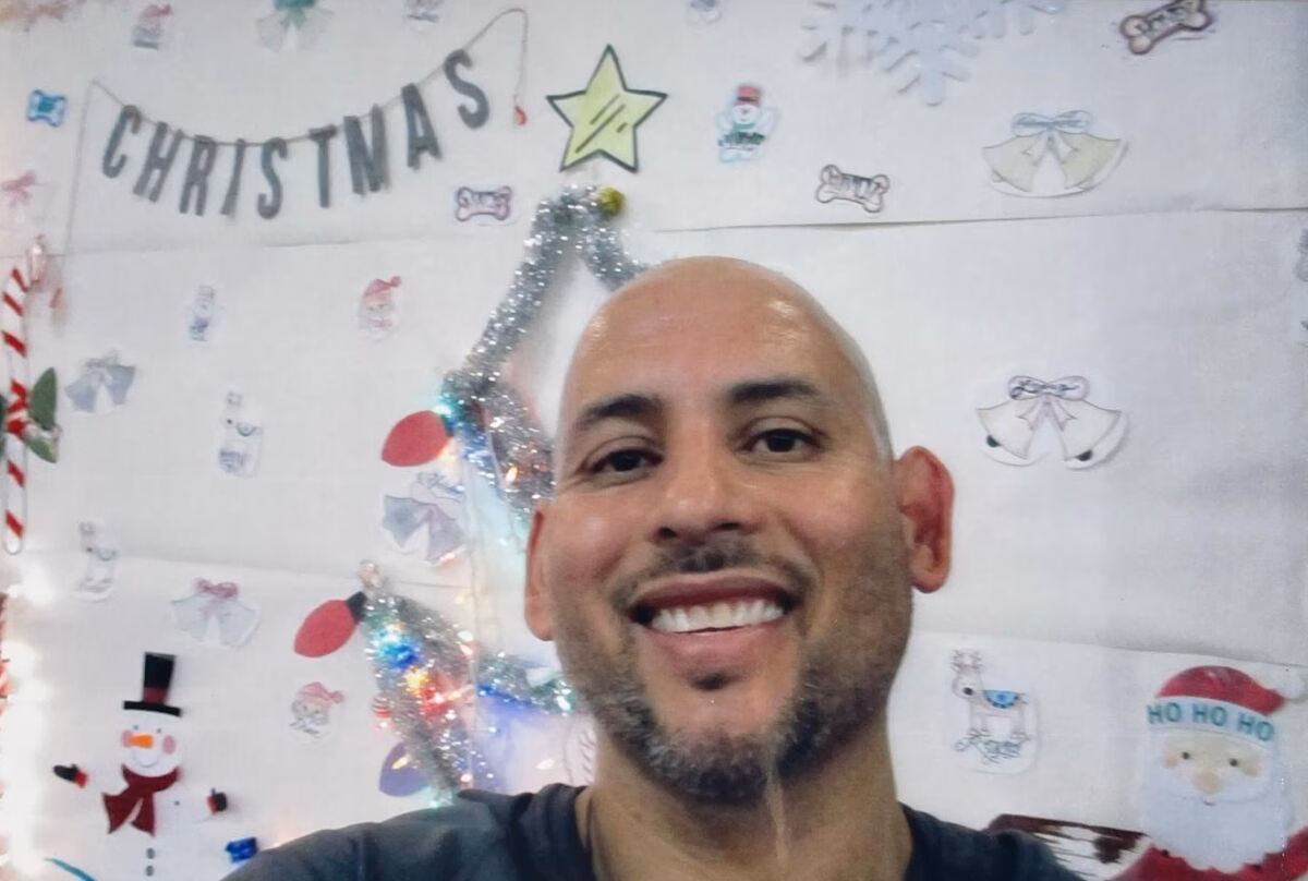A smiling man with a beard in front of a wall decorated with Christmas-themed stickers and a 'CHRISTMAS' banner.