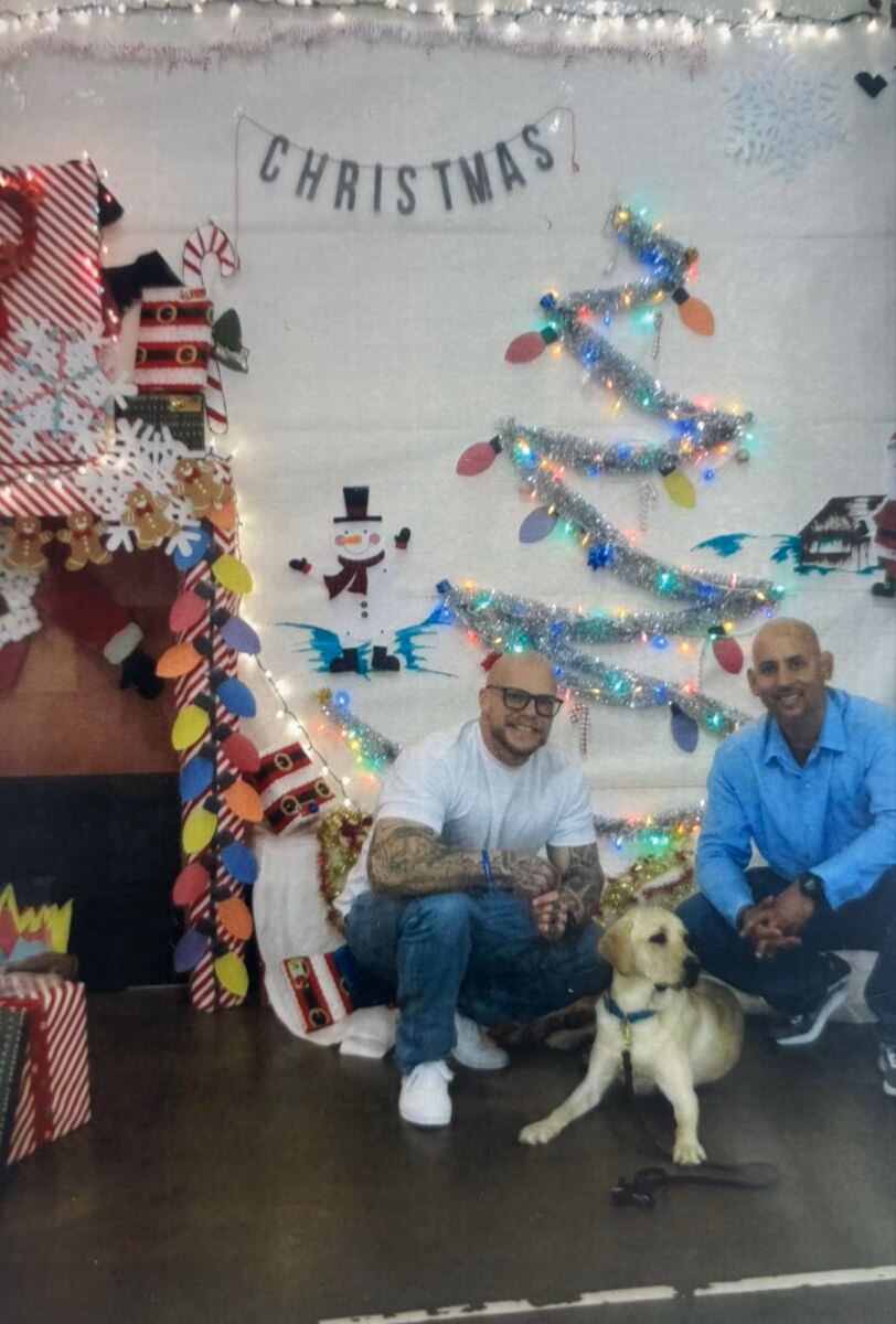 Two men and a yellow Labrador retriever pose in front of a festive Christmas backdrop featuring a tinsel tree, a snowman, and a 'CHRISTMAS' banner.
