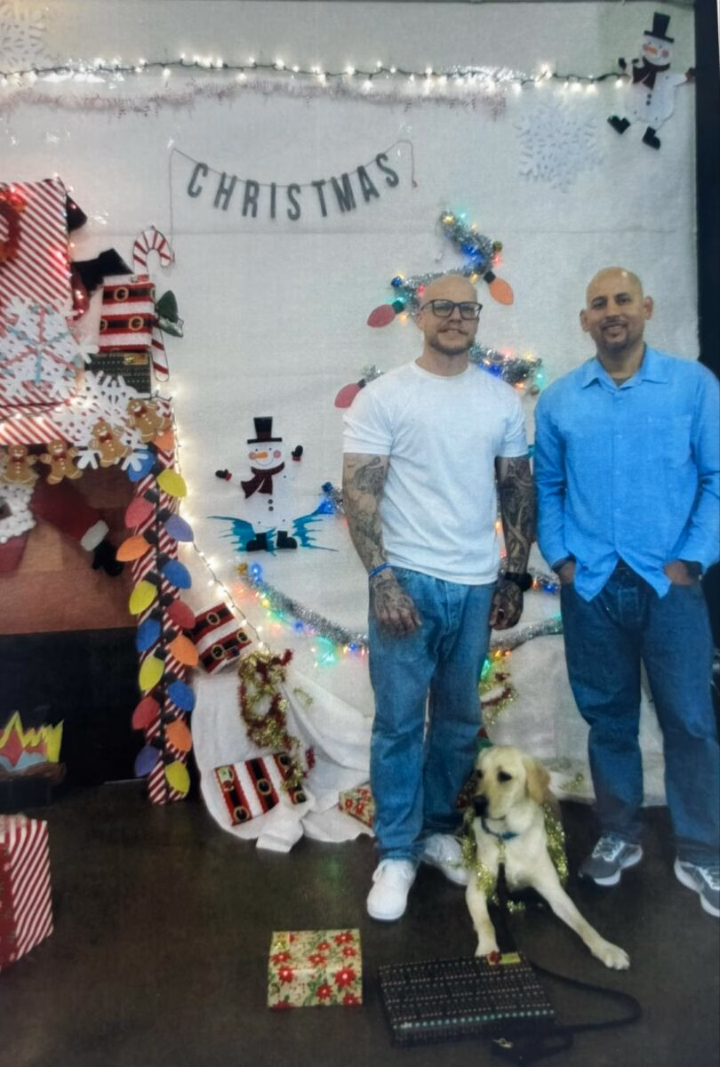 Two men and a golden retriever dog pose in front of a decorated Christmas backdrop featuring a 'CHRISTMAS' banner, snowmen, and lights.