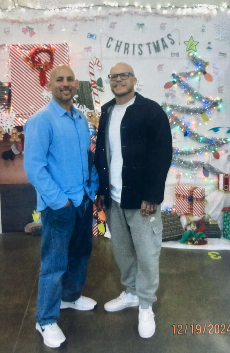 Two men, Eric and a companion, stand smiling in front of a festive Christmas backdrop decorated with lights, a tree, and a banner.